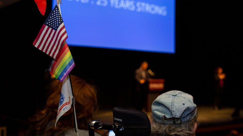 person in wheel chair with flags on back watches speaker on stage