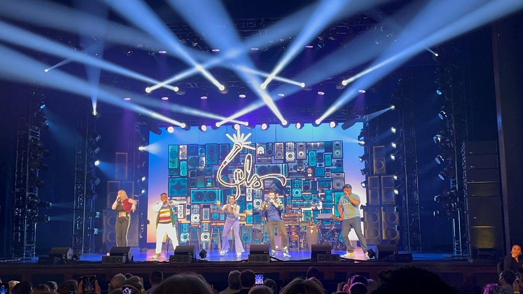 Cell phone showing a theater stage with five people on it holding mics with white lights surrounding the stage shinging on the audience as a performance of Freestyle Love Supreme in Washington D.C.