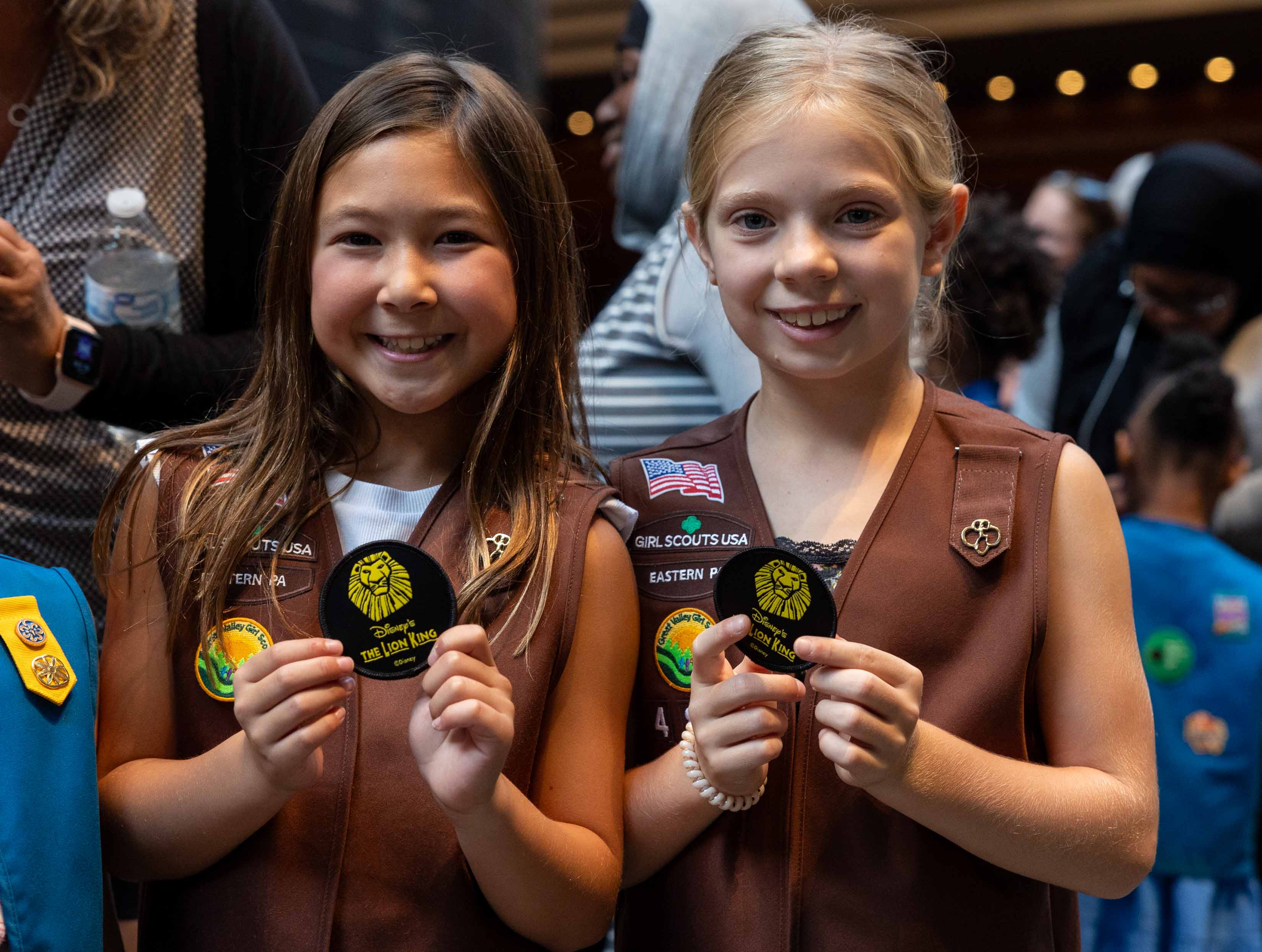 Two young girls in Gril Scout vests hold small badges embroidered with Disney's The Lion King logo