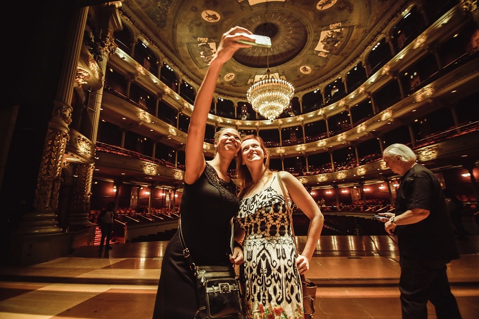Two people pose for a selfie with a phone on stage of a theater