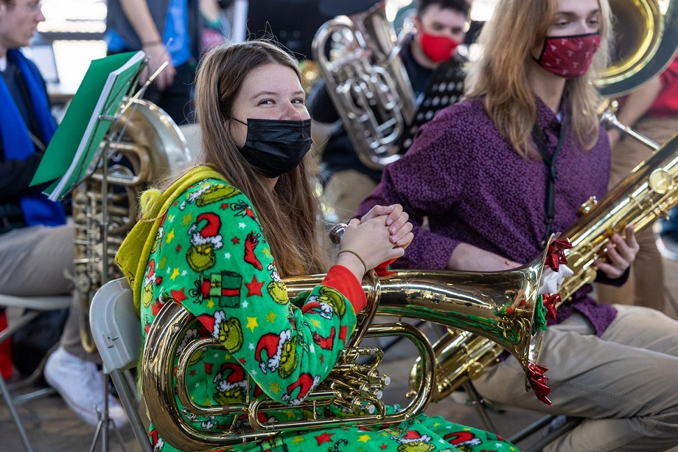 A young person in Christmas attire sits and plays a tuba among a group