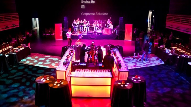 Guests dance and chat as a live band plays music and bartenders serve drinks.